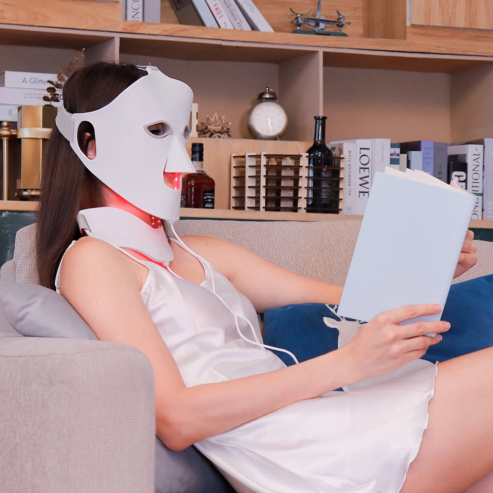 A person wearing a flexible, white silicone LED face and neck mask with red light therapy, while reading on a sofa.
