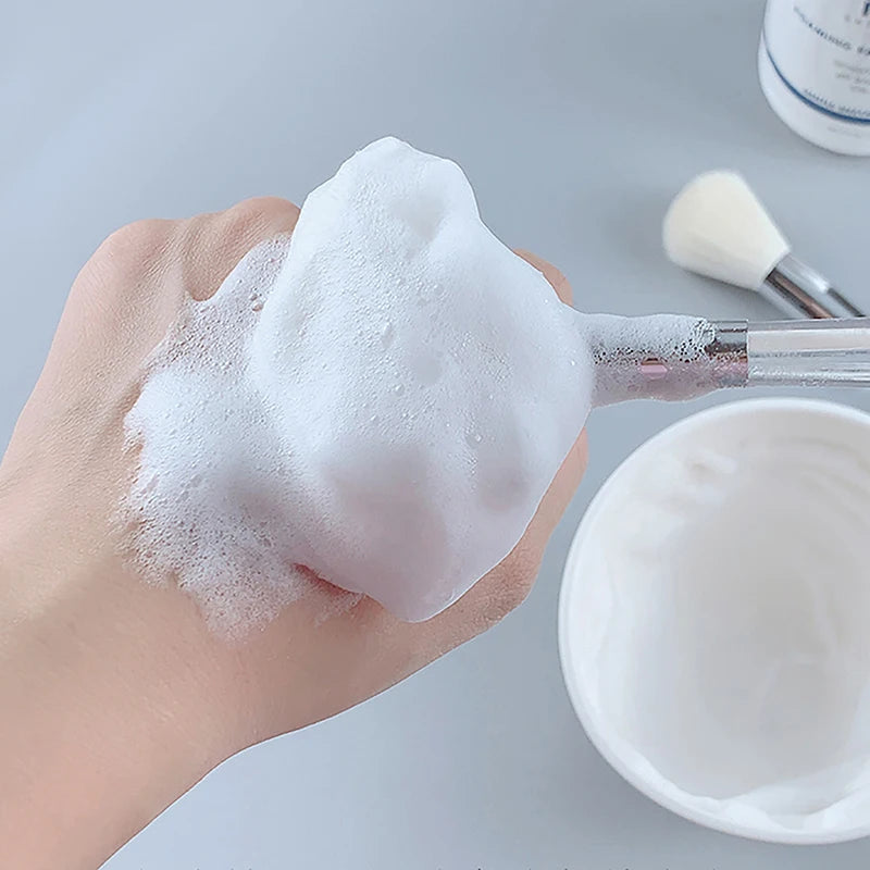 Soft facial cleanser brush applying foamy skin care product to hand, portable cosmetic tool with white bristles.