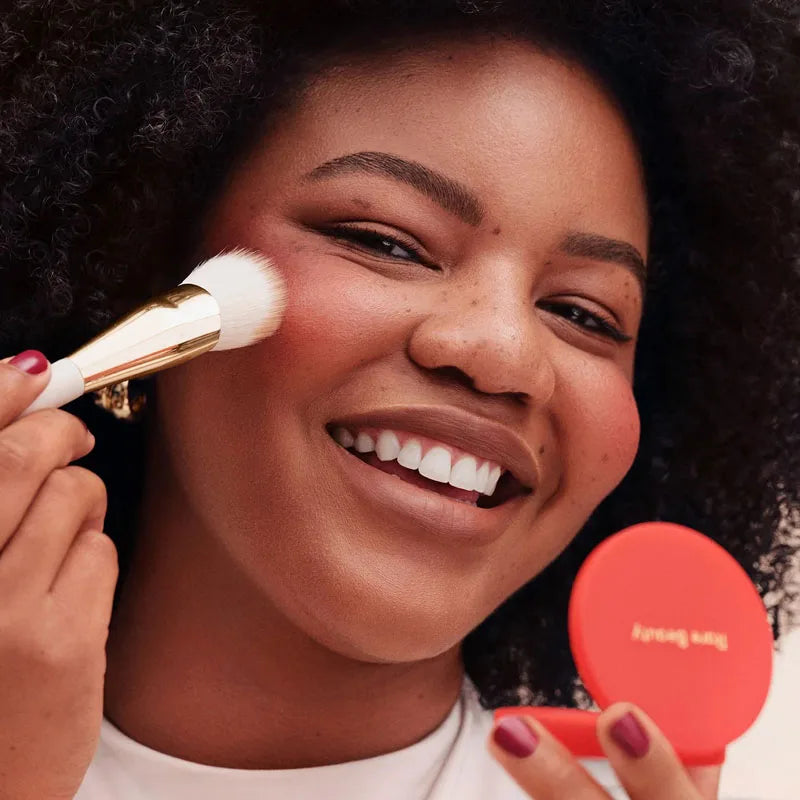 Rare Beauty powder blush with smooth, blendable texture being applied with a gold brush for a natural, rosy glow.