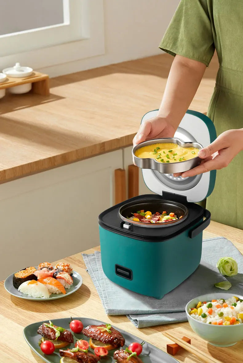 Mini electric rice cooker on countertop with open lid and steaming insert, teal body.