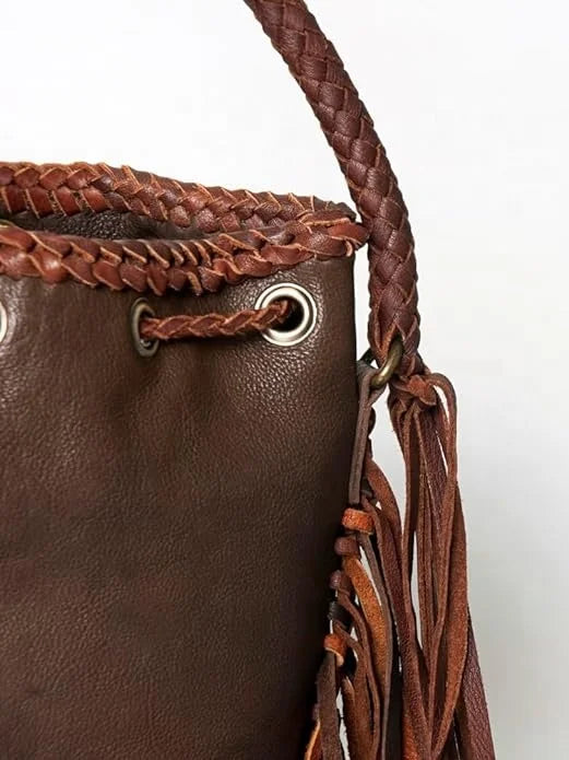 Brown vintage leather bucket bag with braided trim, metal eyelets, and boho fringe detail on a woven shoulder strap.