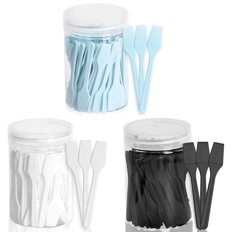 Three clear jars filled with mini blue, white, and black plastic cosmetic spatulas for makeup and skincare application.