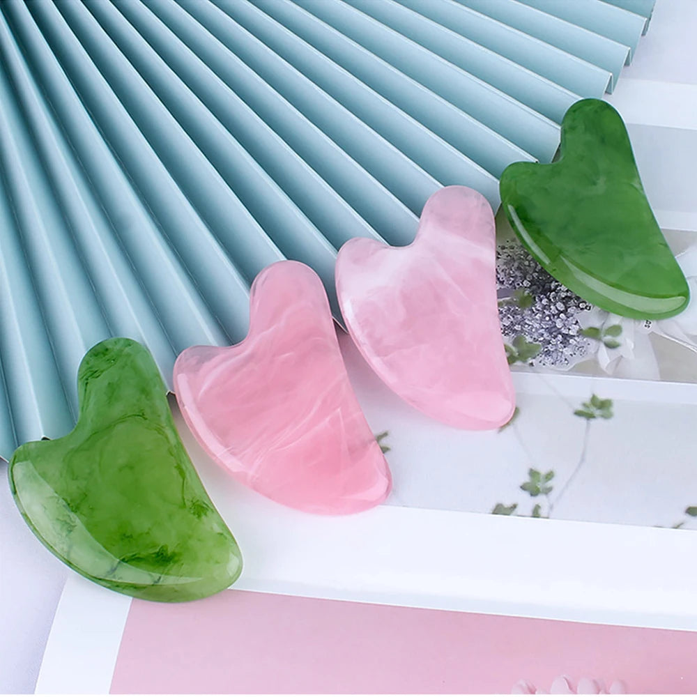 Two green and two pink heart-shaped resin gua sha massage tools on a white surface with a blue pleated background.