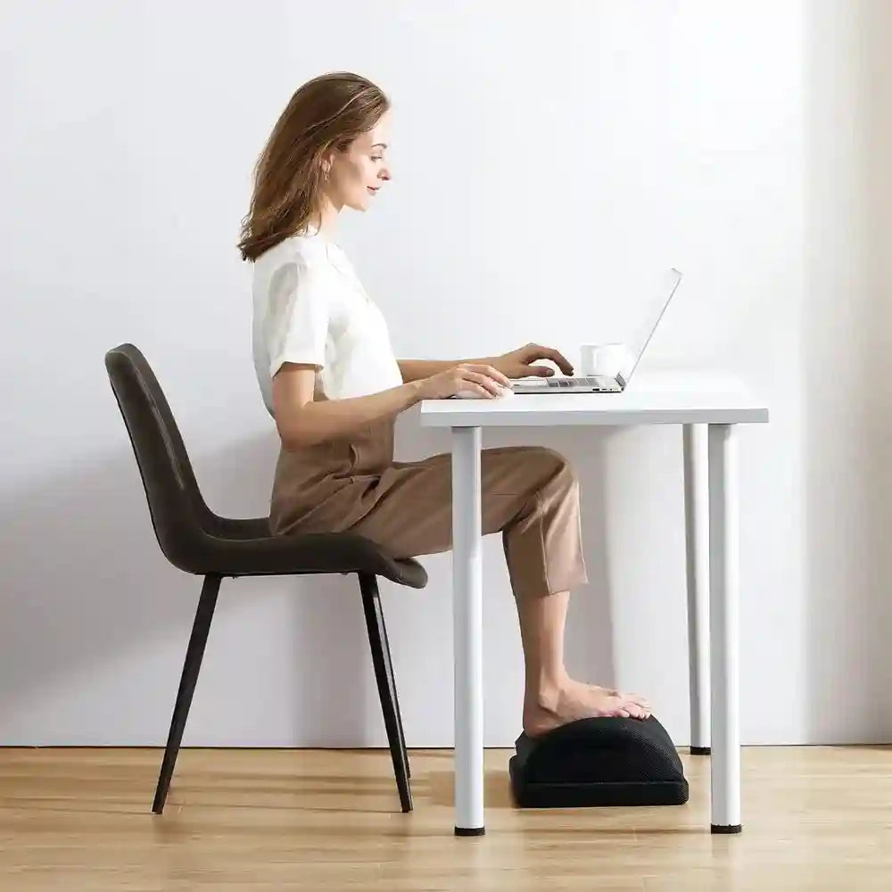 Woman using black memory foam under desk foot rest for ergonomic office posture and pain relief.