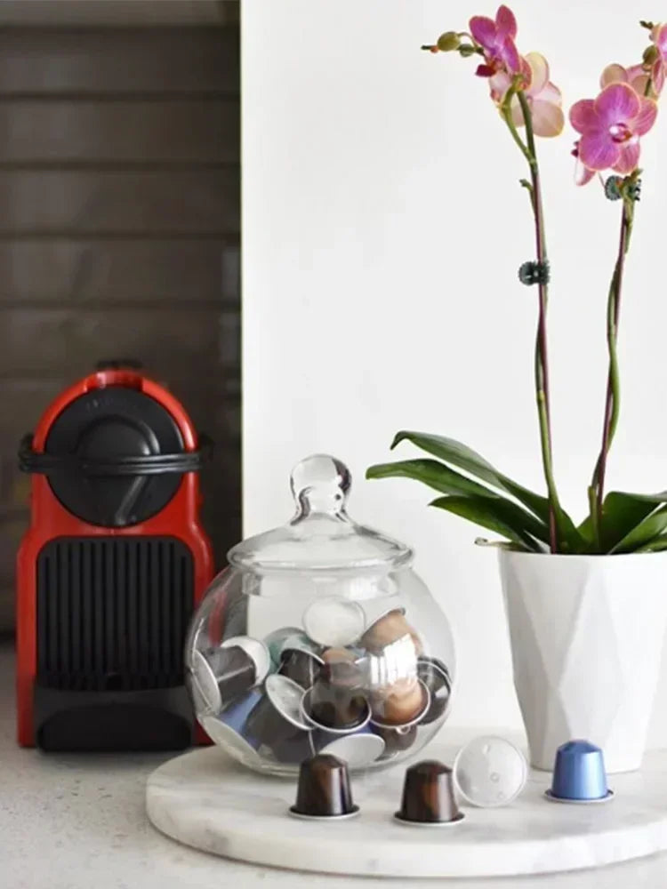 Red Nespresso coffee machine with glass capsule jar and coffee pods on a white marble surface.