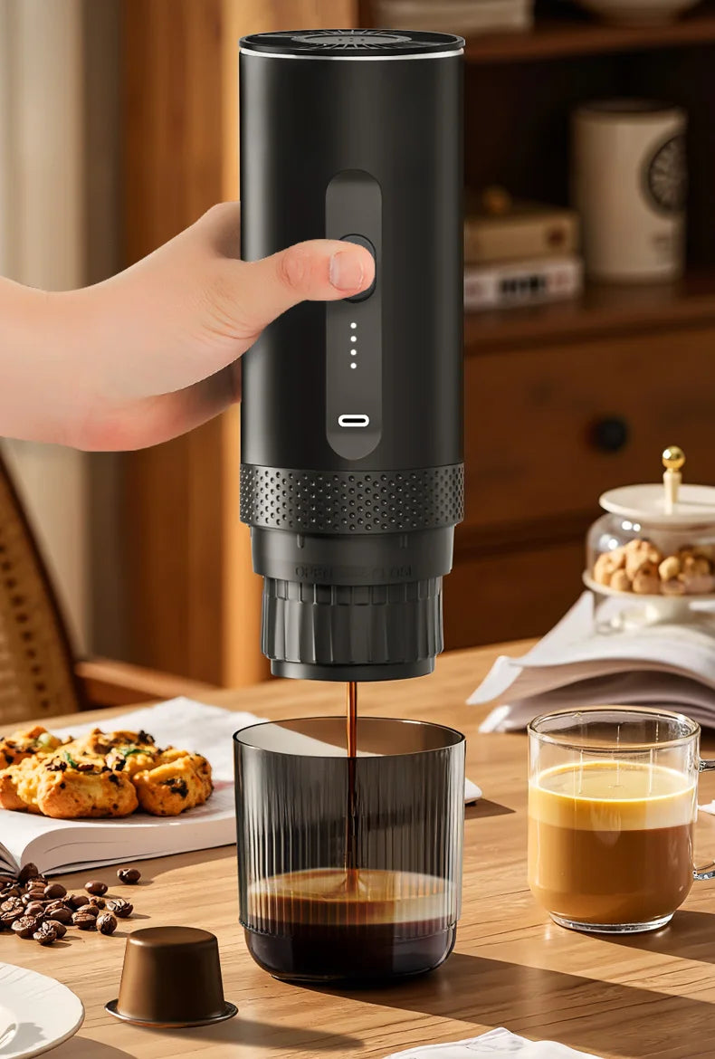 Handheld black portable electric espresso coffee maker brewing into a glass cup on a wooden table.