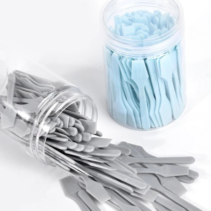 Two clear jars filled with many small, light blue and grey plastic cosmetic spatulas for applying creams and masks.