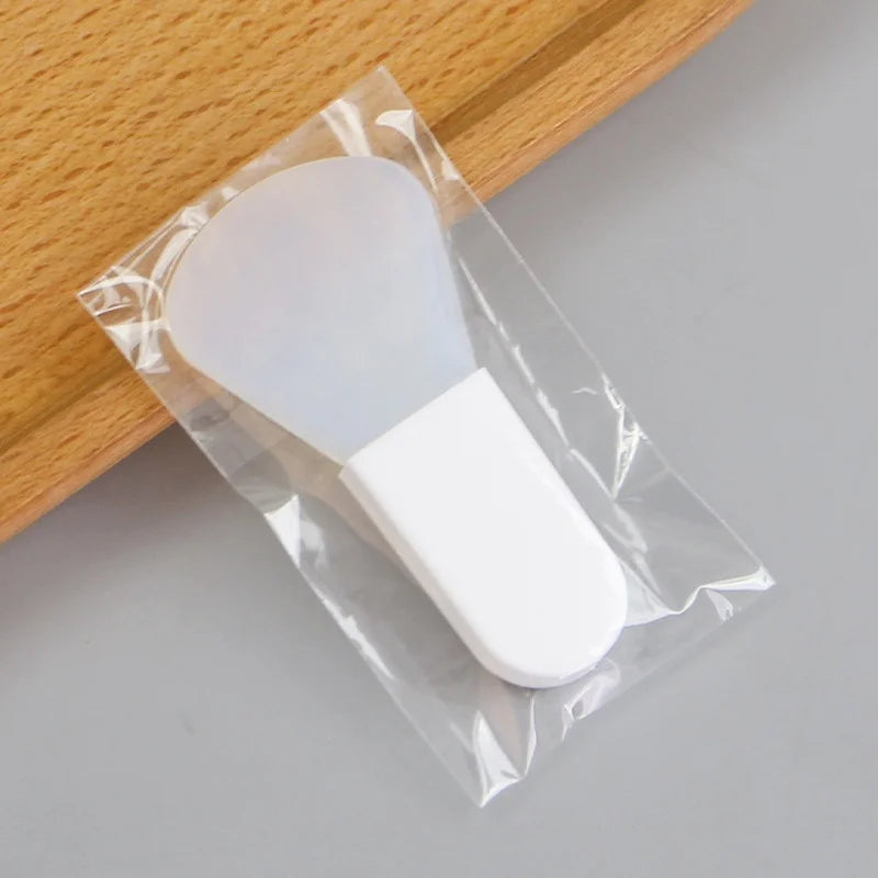 A white silicone facial mask applicator tool with a wide paddle head, sealed in a clear plastic bag.