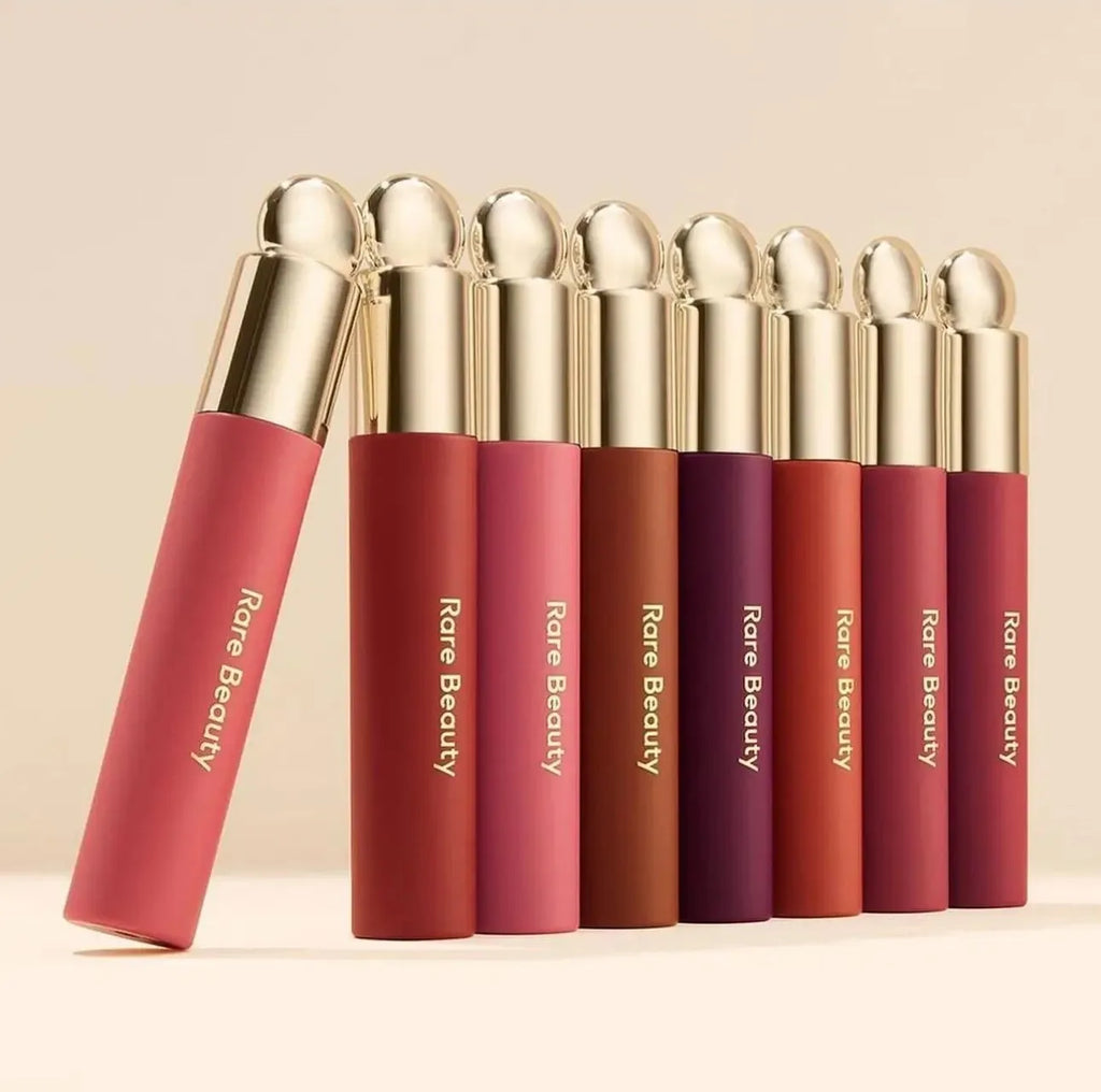 Rare Beauty lip makeup collection with matte finish tubes in various shades and metallic gold caps on beige background.