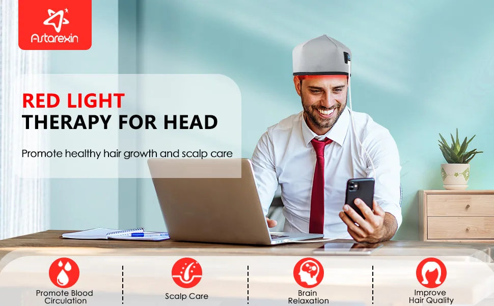Man wearing gray wired LLLT red light therapy cap for hair growth at desk with laptop and phone.