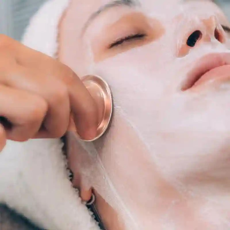 A person using a rose-gold facial cupping tool on skin covered in white cream for an anti-wrinkle massage.