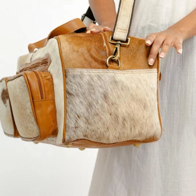 Western-style brown leather and hair-on-hide travel duffel trunk bag with side zip pockets and shoulder strap.