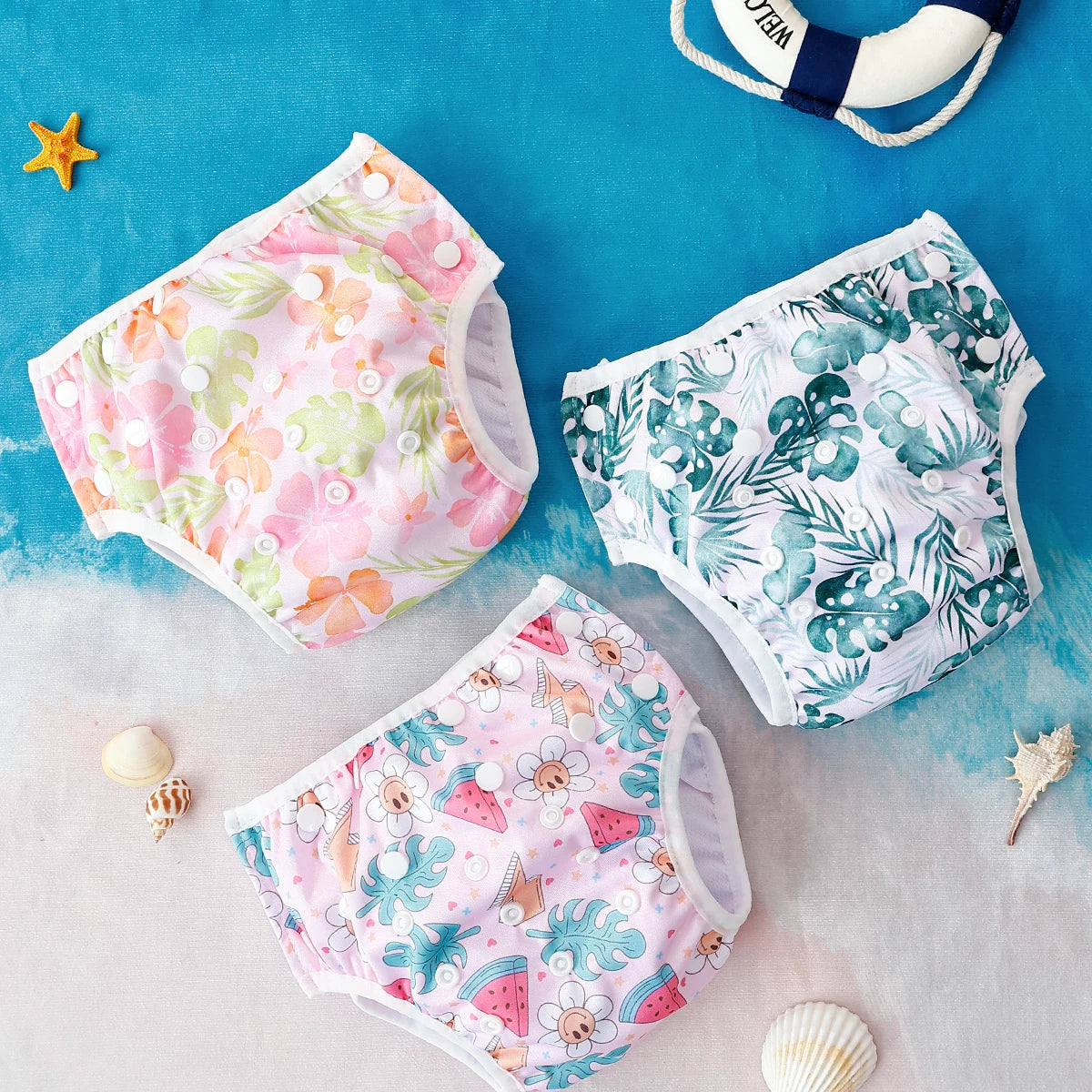Three reusable baby swim diapers with tropical, floral, and watermelon prints on a beach background.
