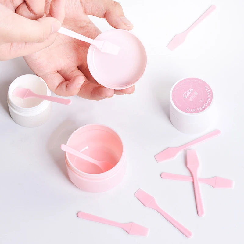 A hand holds a small pink cosmetic spatula above pink and white jars, surrounded by several scattered pink spatulas.