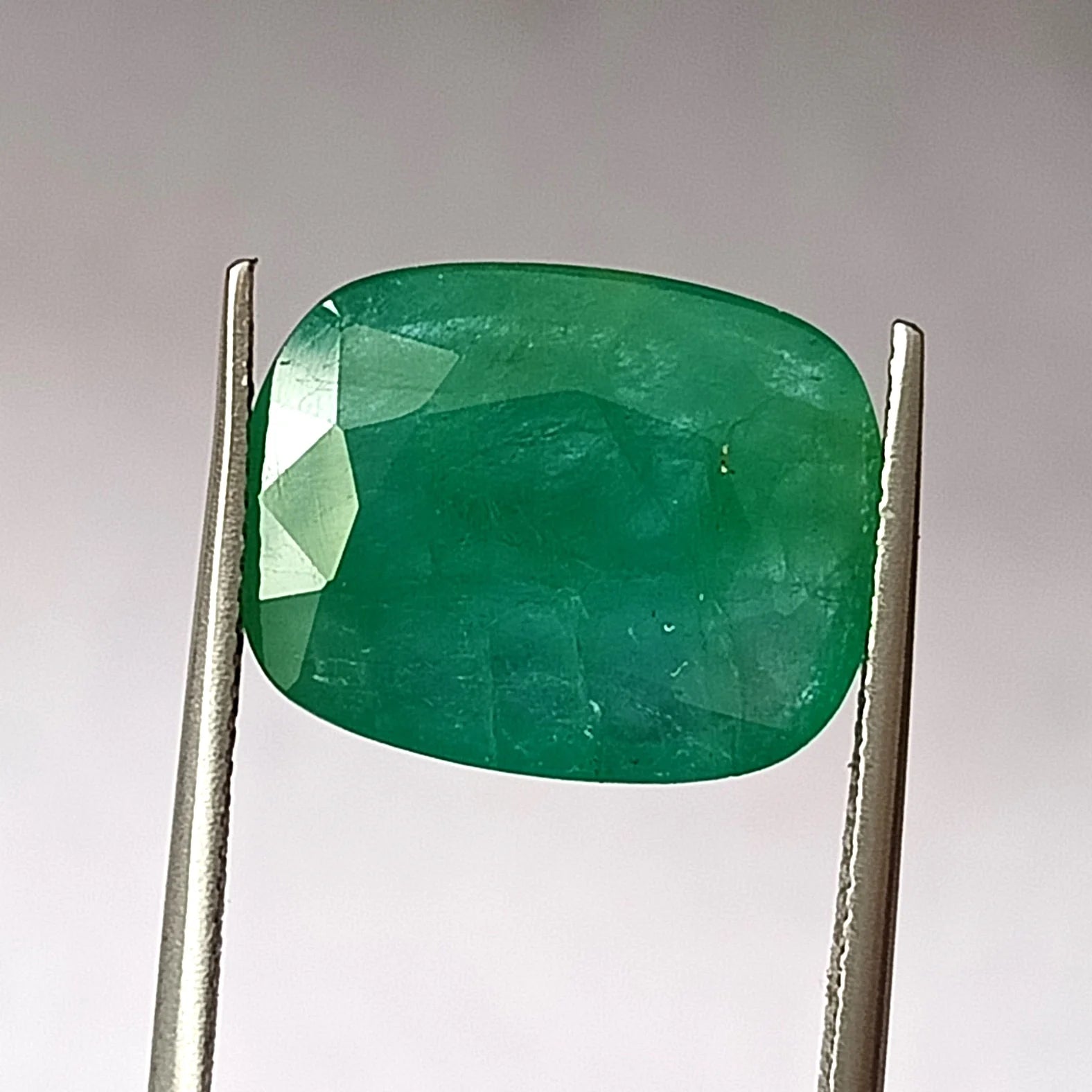 Green oval emerald gemstone, faceted, front view in metal tweezers with bright facet.