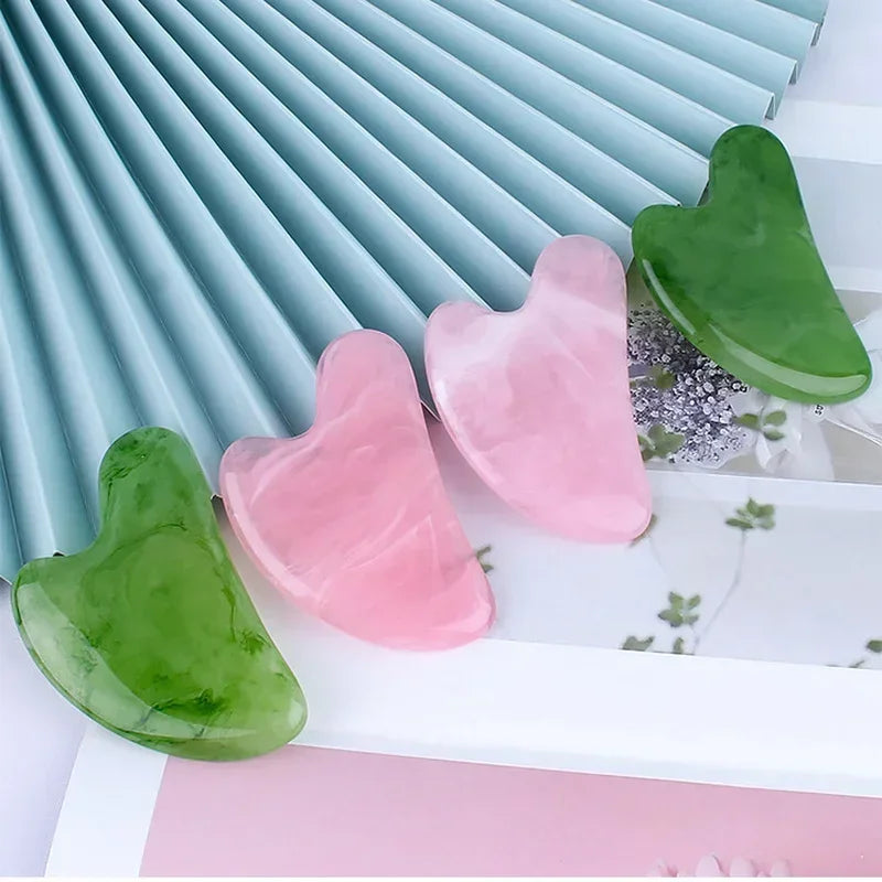 Four heart-shaped resin gua sha massage tools in shades of green and pink, arranged on a decorative fan background.