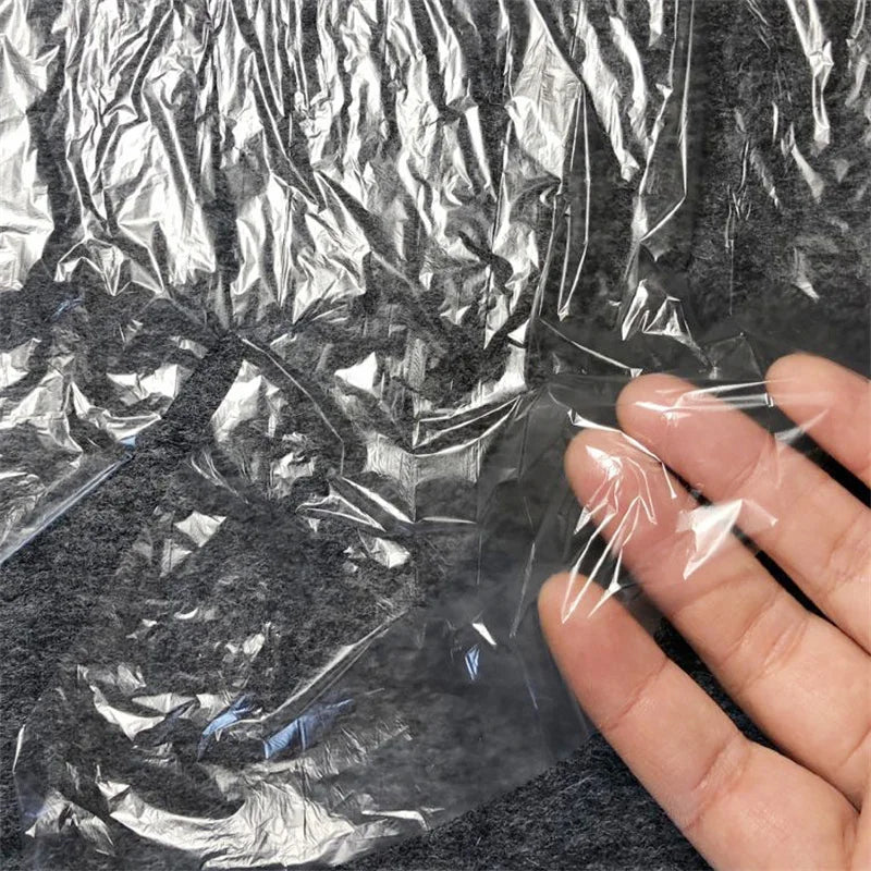 A hand holds a thin, transparent, crinkled plastic film sheet against a dark gray textured background.