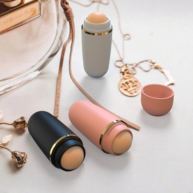 Three oil-absorbing volcanic stone rollers in matte black, pink, and grey with gold trim, arranged on a table.