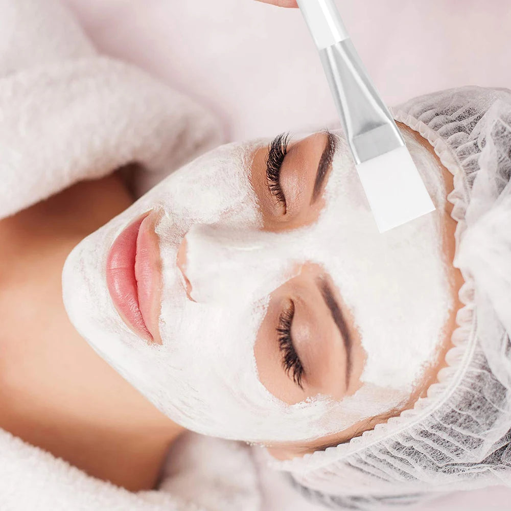 A white flat-bristled facial mask brush applying a white cream mask onto a woman’s face in a spa setting.