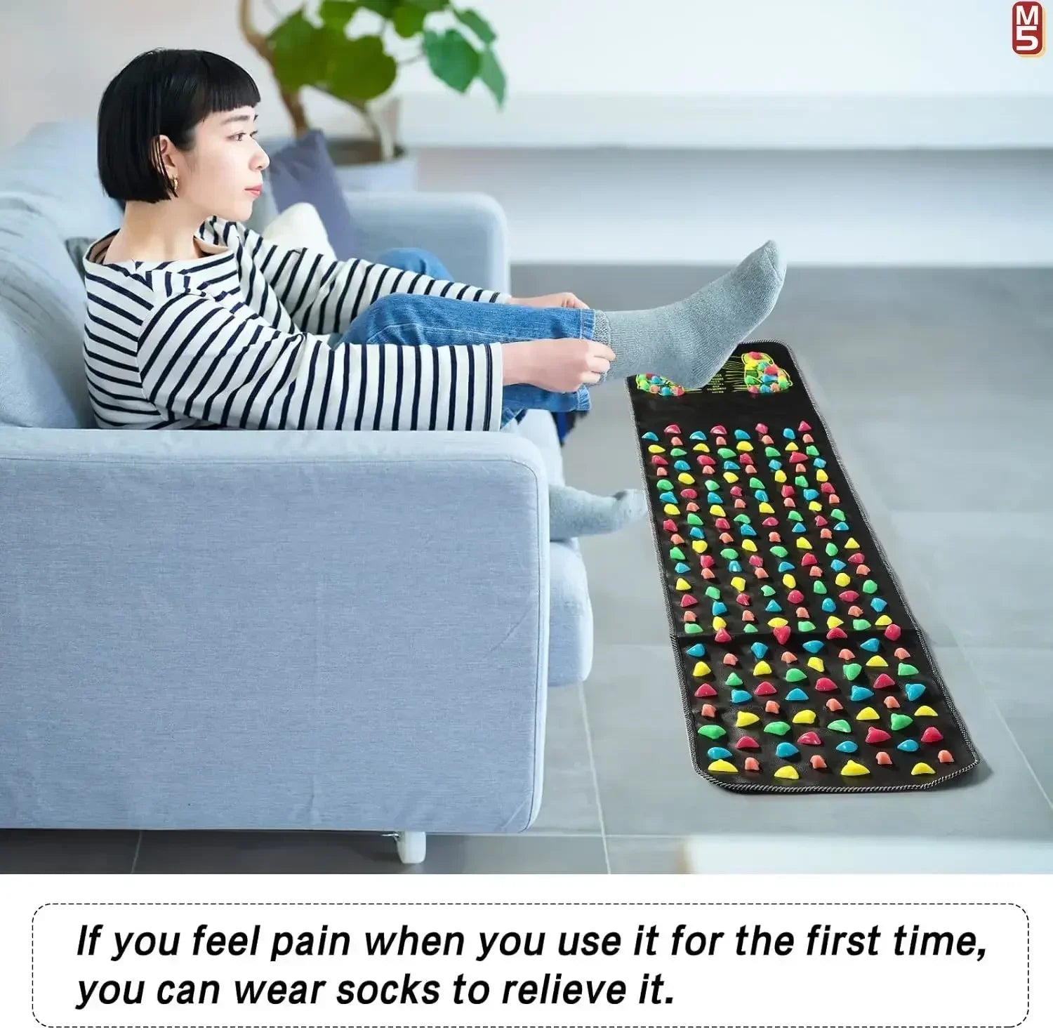 Woman using black acupressure foot reflexology massage pad with colorful nodes while sitting on a couch.
