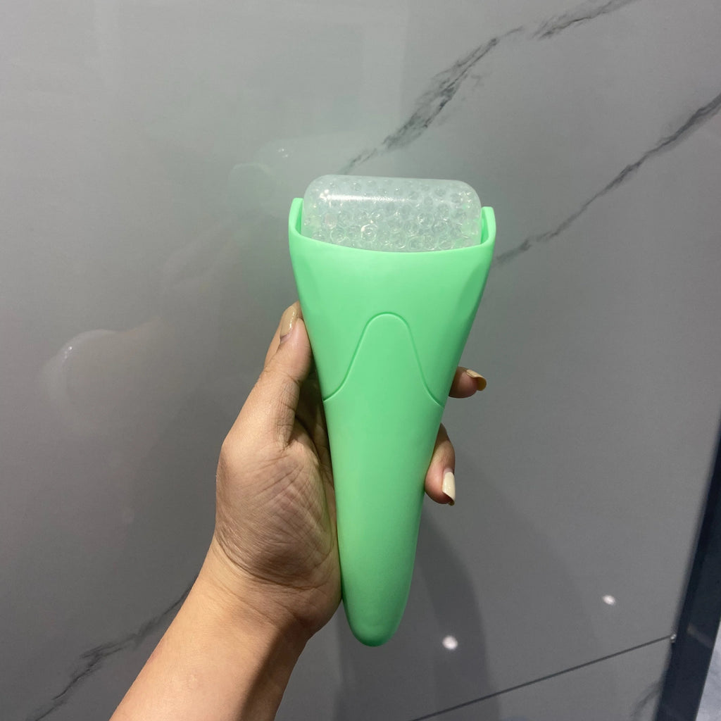 A hand holds a mint green facial ice roller with a textured, bumpy translucent barrel against a marble background.