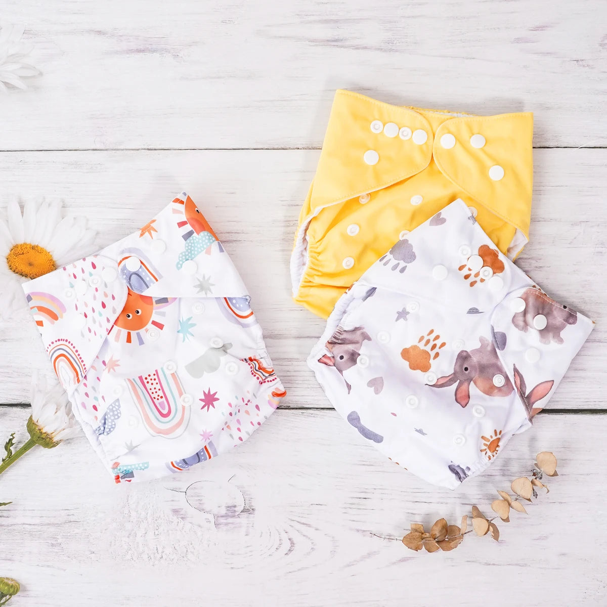 Three reusable cloth baby diapers featuring yellow, rainbow, and bunny patterns on a rustic wooden background.
