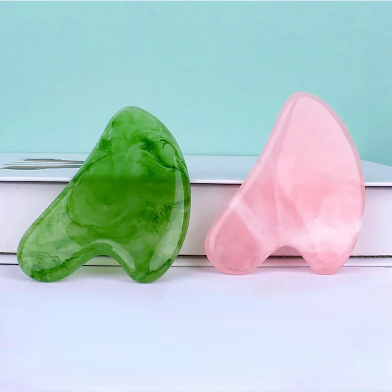 Two smooth, wing-shaped resin gua sha facial massagers in vibrant green and pale pink marble-effect patterns.