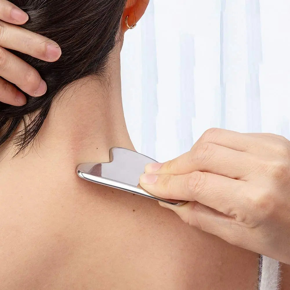 A hand uses a smooth, heart-shaped stainless steel gua sha tool to massage the back of a person's neck.