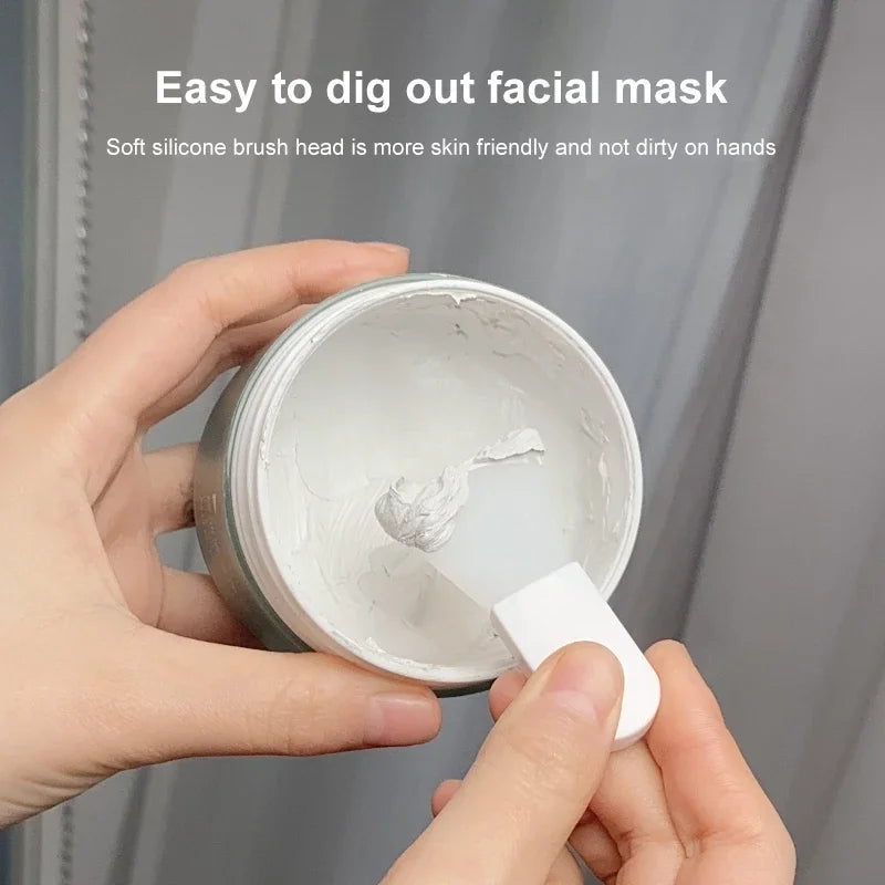 A person uses a small, white, flexible silicone applicator to scoop white facial mask from a circular jar.