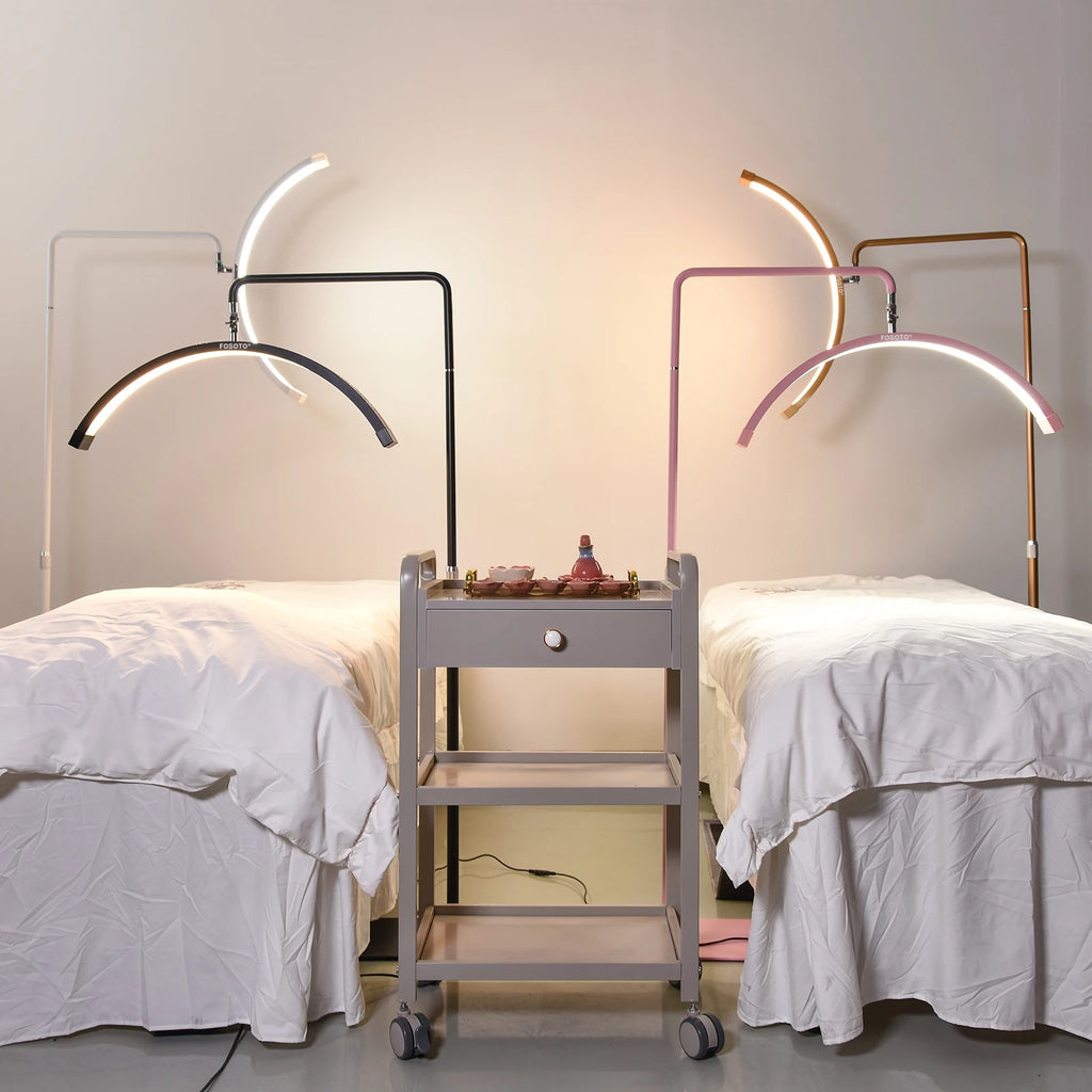 Two curved LED lash lamps on adjustable stands over beauty beds, flanking a grey three-tier rolling salon cart.