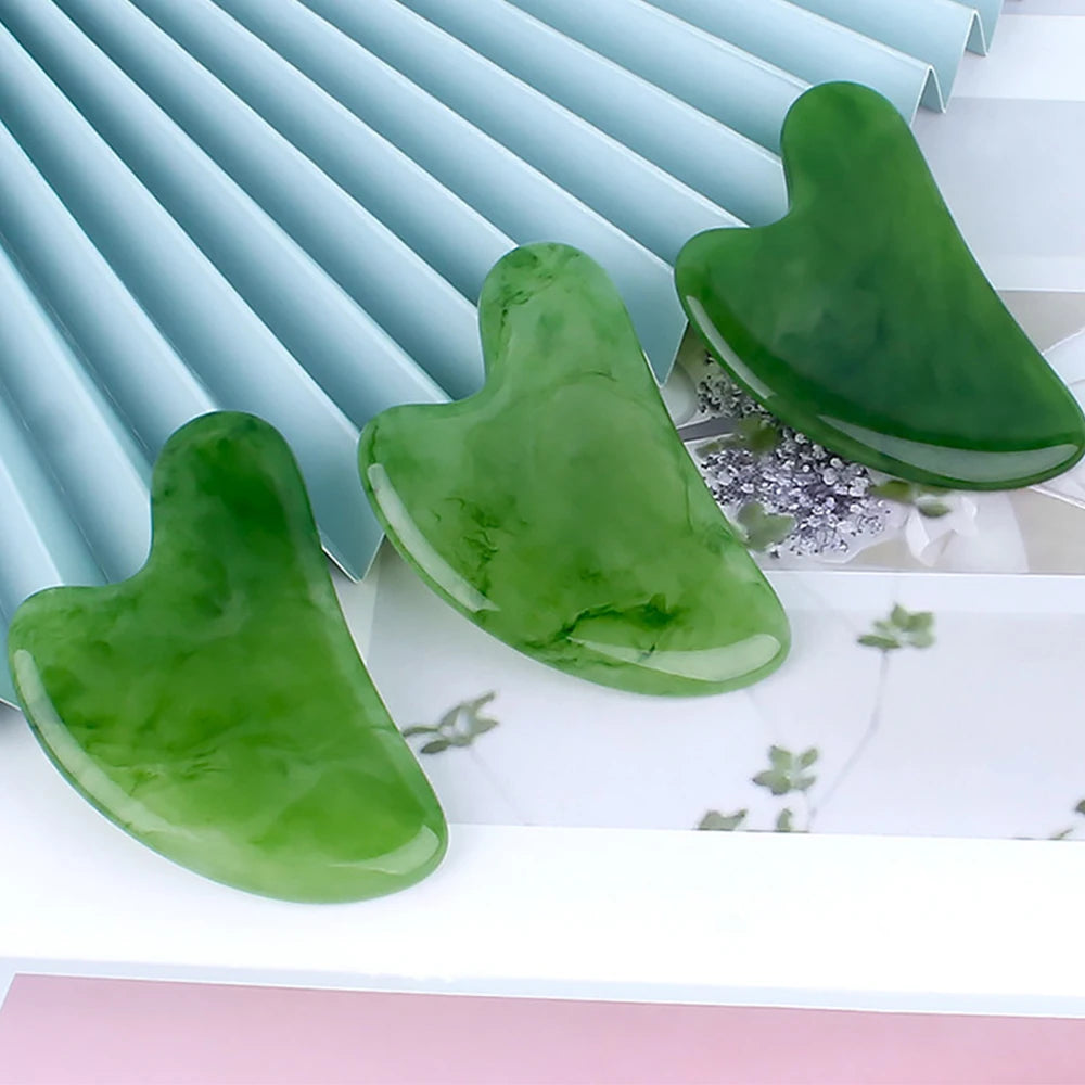 Three heart-shaped green resin gua sha massage tools displayed against a blue fan and floral background.