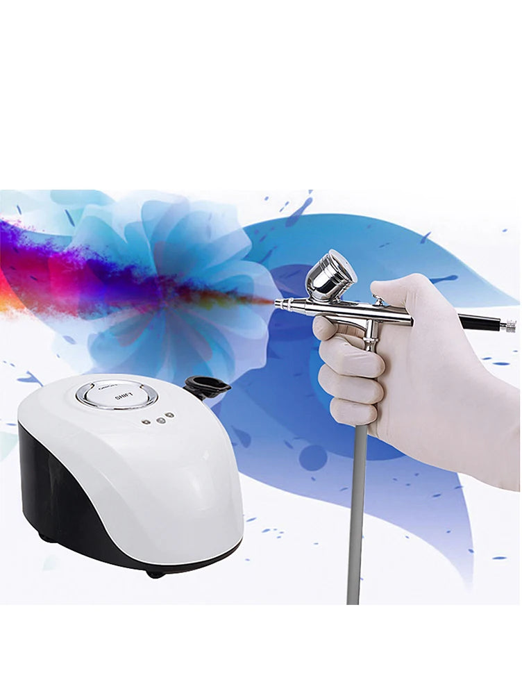 A gloved hand holds a chrome airbrush connected to a white and black tabletop air compressor machine.