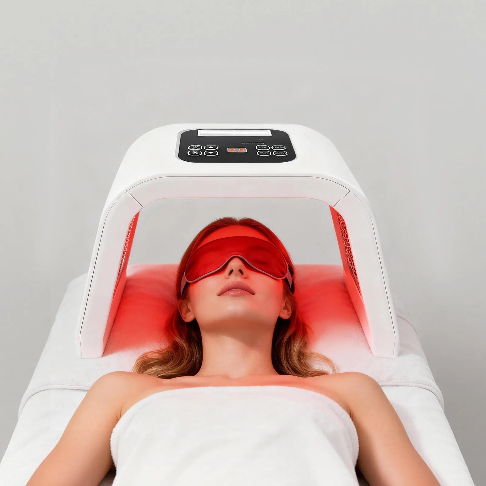 A person wearing red goggles lies under a white, arched LED light therapy machine emitting red light for skincare.