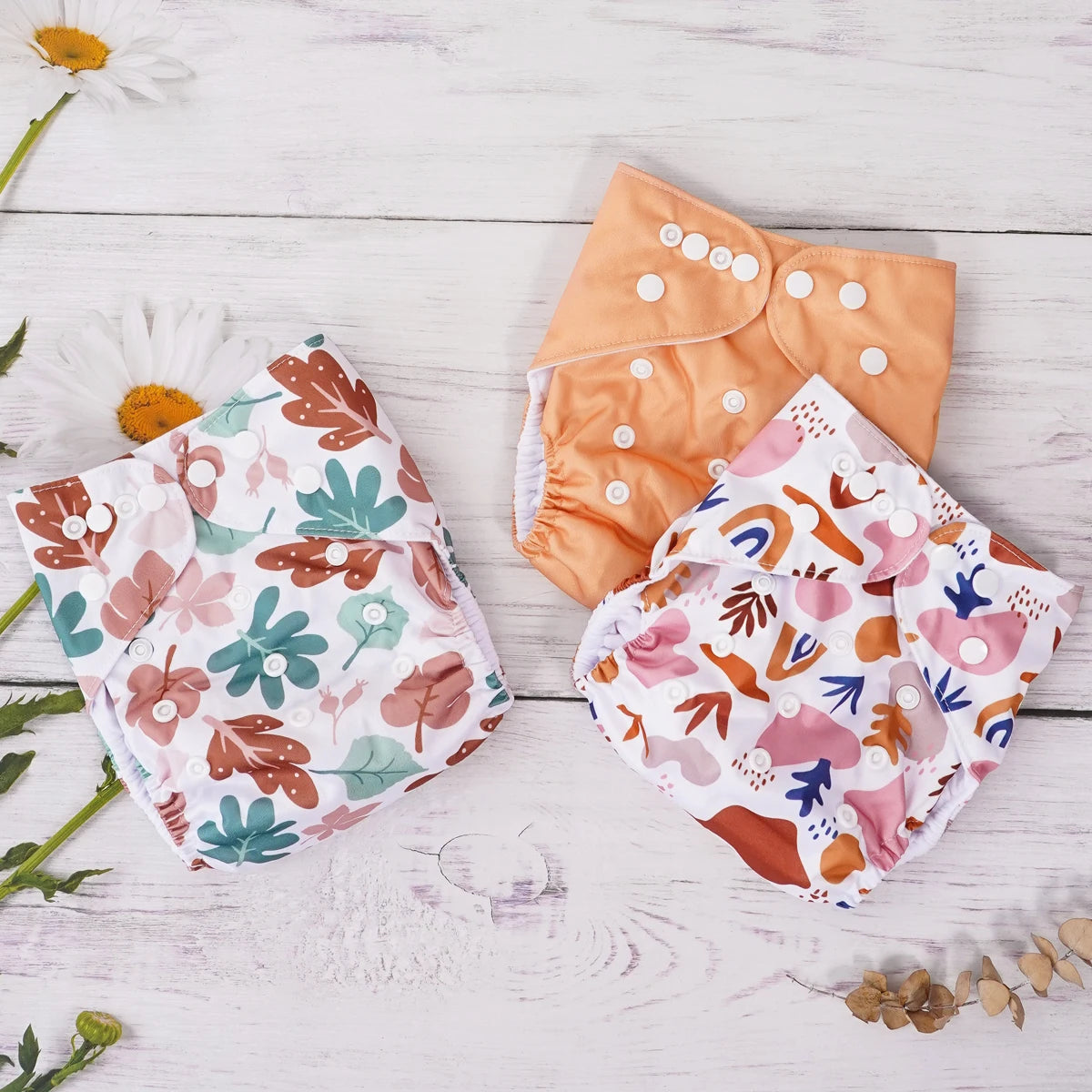 Three cloth baby diapers with adjustable snaps, featuring leaf and abstract patterns on a white wooden background.
