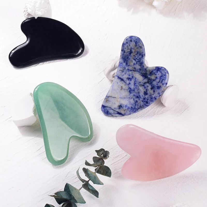 Four heart-shaped stone Gua Sha facial massage tools in black, green, blue-speckled, and pink on a white surface.
