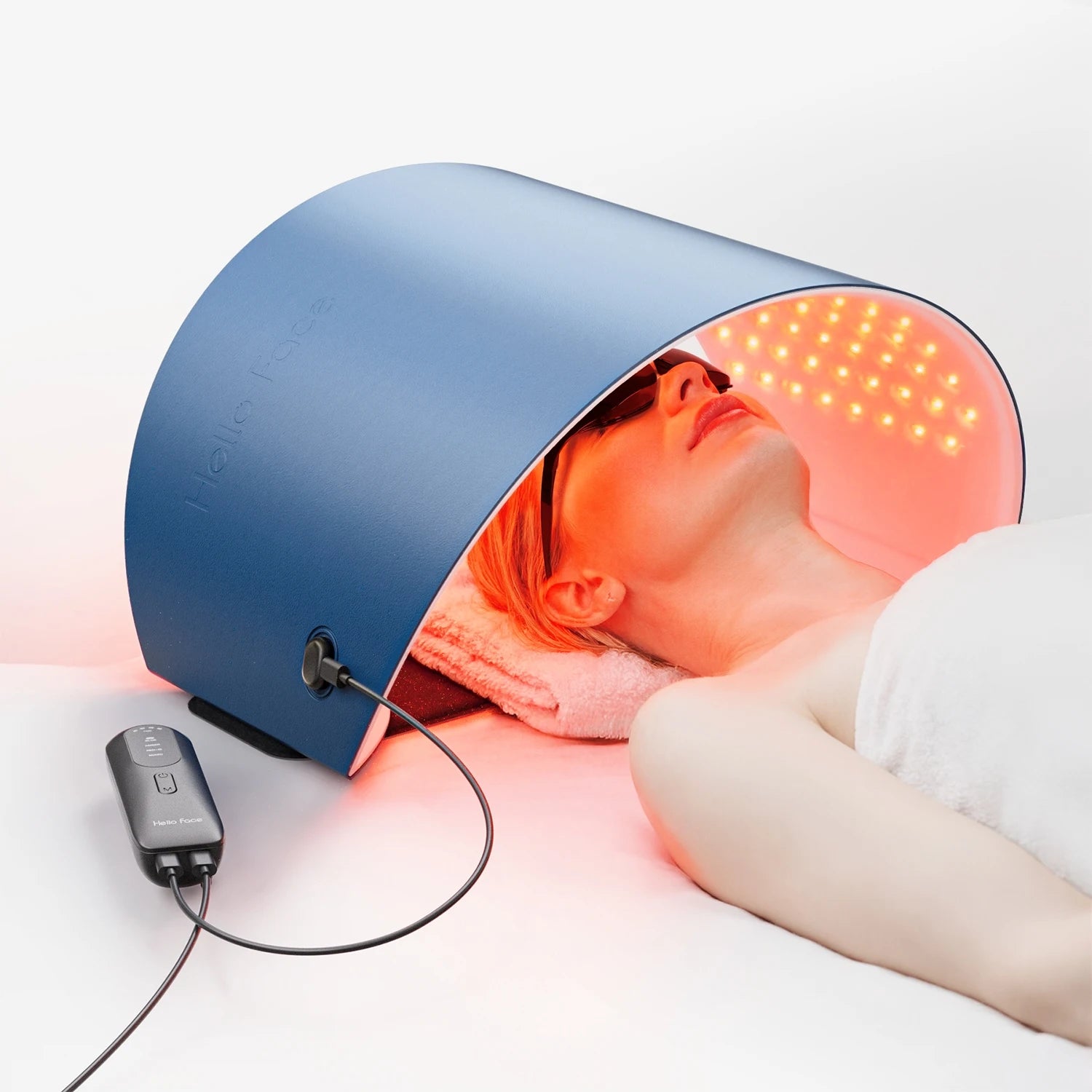 A woman lies under a curved blue Hello Face LED light therapy mask emitting red light, connected to a handheld remote.