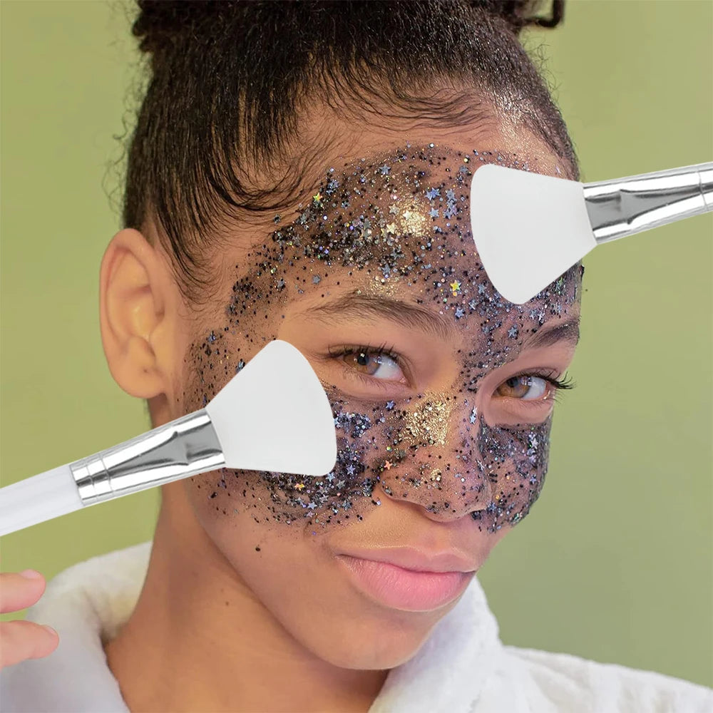 Two white, soft-tip silicone face mask brushes with silver ferrules being used on a person wearing a sparkly face mask.
