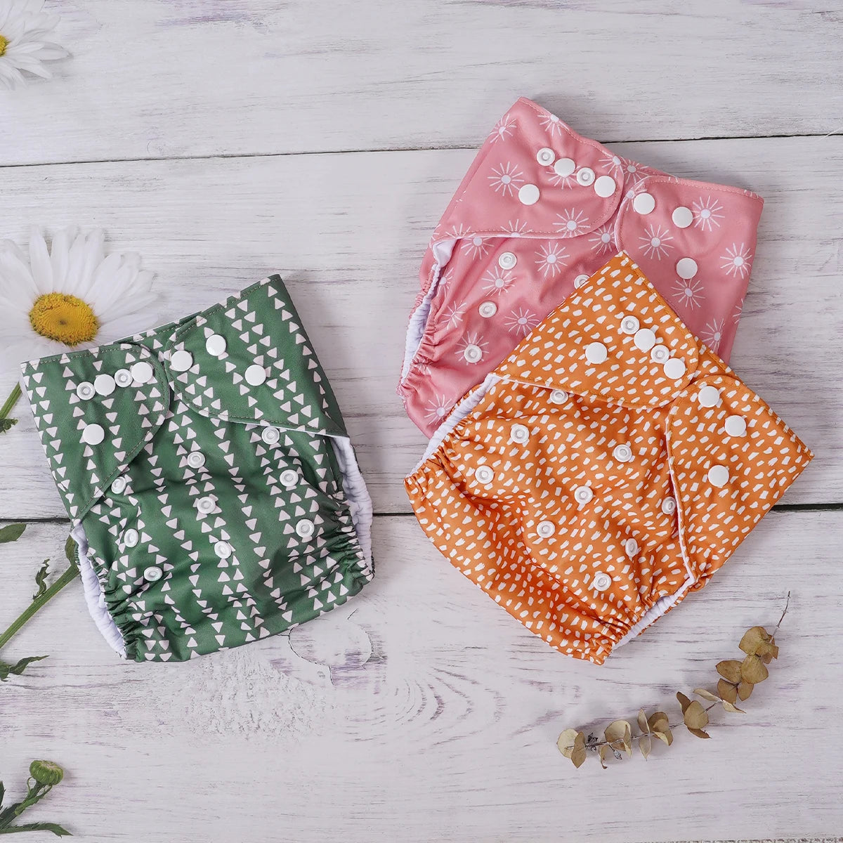 Three reusable cloth baby diapers in green, pink, and orange patterns with adjustable snap buttons.