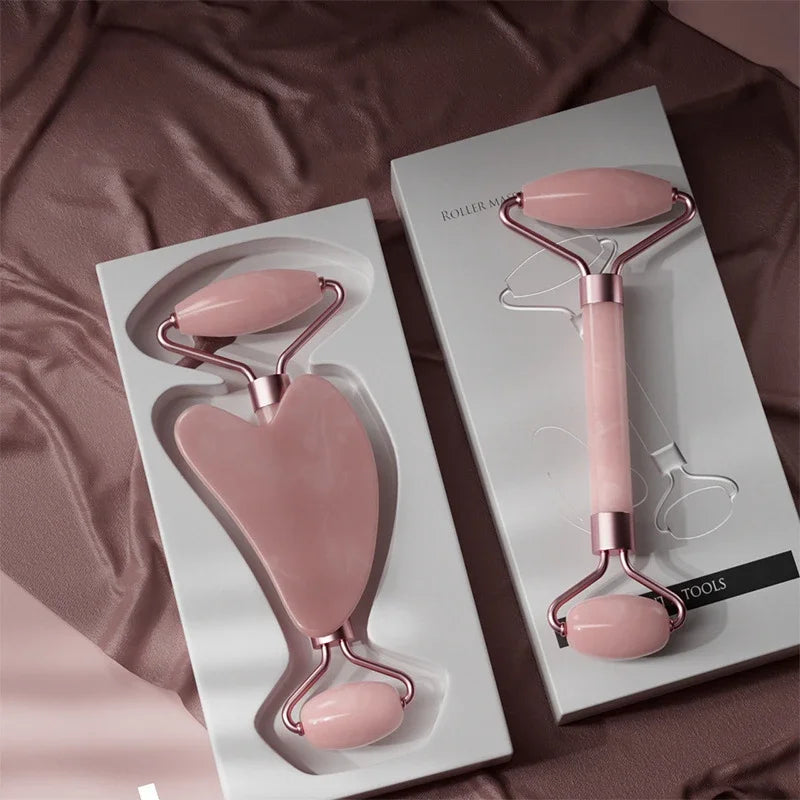 Two pink resin facial massagers—a dual-ended roller and a heart-shaped gua sha tool—in white packaging on silk fabric.