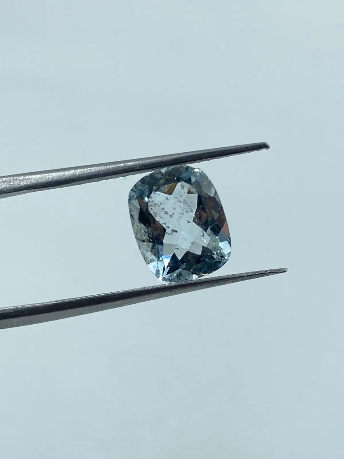 Loose cushion-cut aquamarine gemstone, sky blue, angled between tweezers, close view