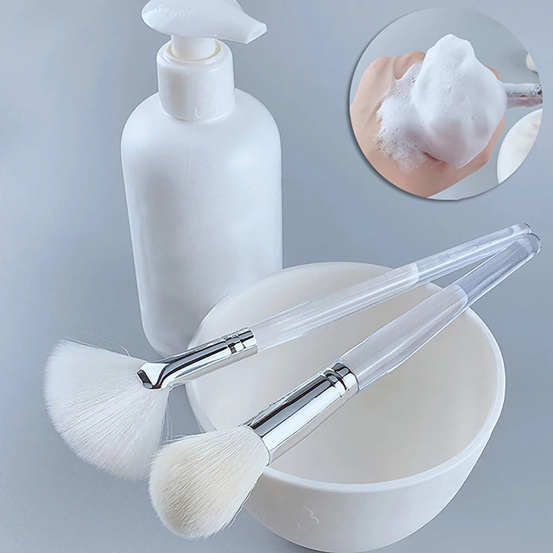 Soft facial cleansing and mask brushes with clear handles, white mixing bowl, and soap dispenser for skin care.