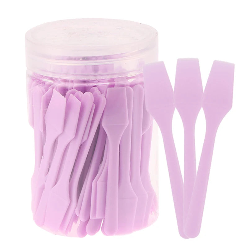 Clear container filled with 100 mini, light purple plastic cosmetic spatulas for makeup and skincare application.
