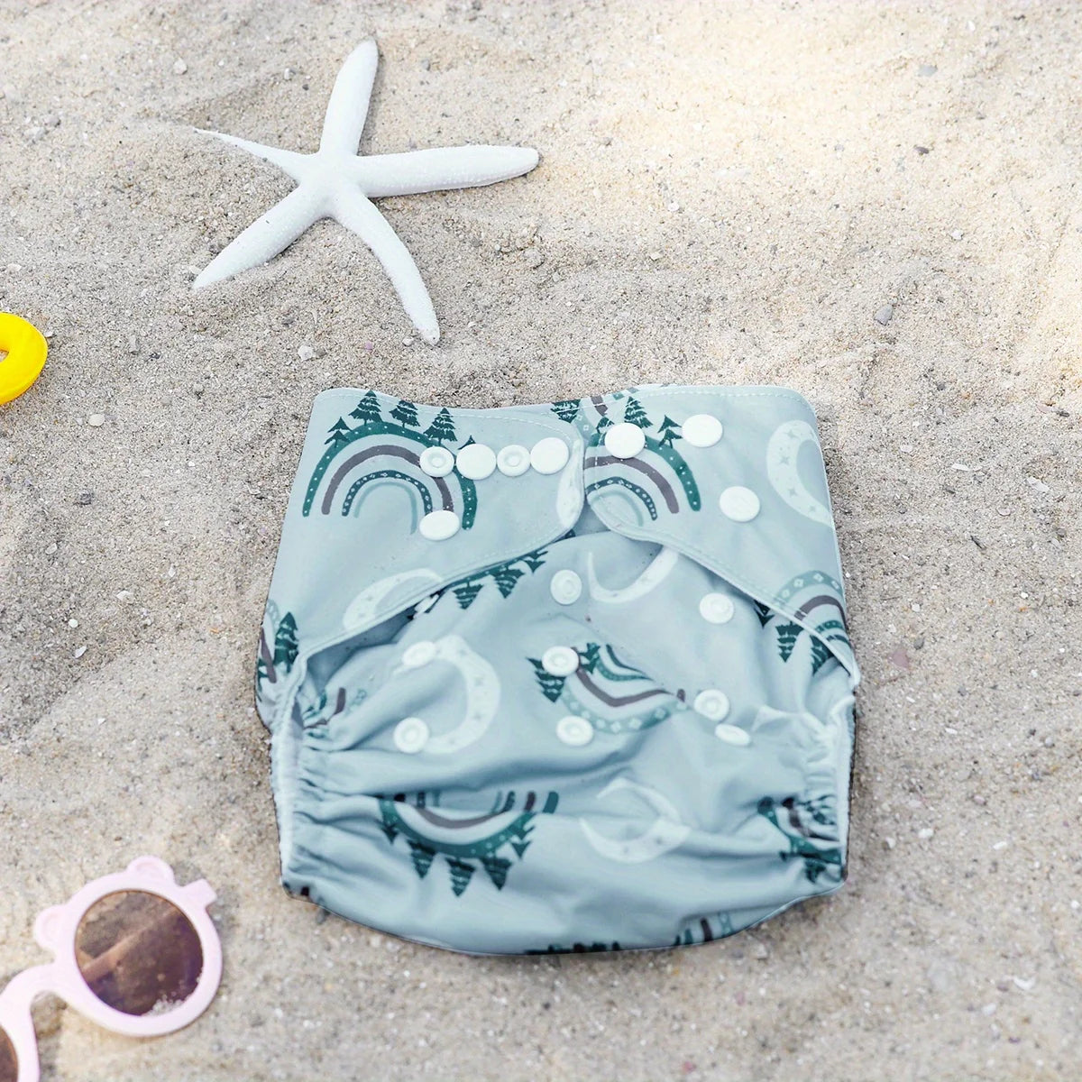 Reusable cloth diaper with mountain rainbow print on sandy beach, perfect for eco-friendly baby care.