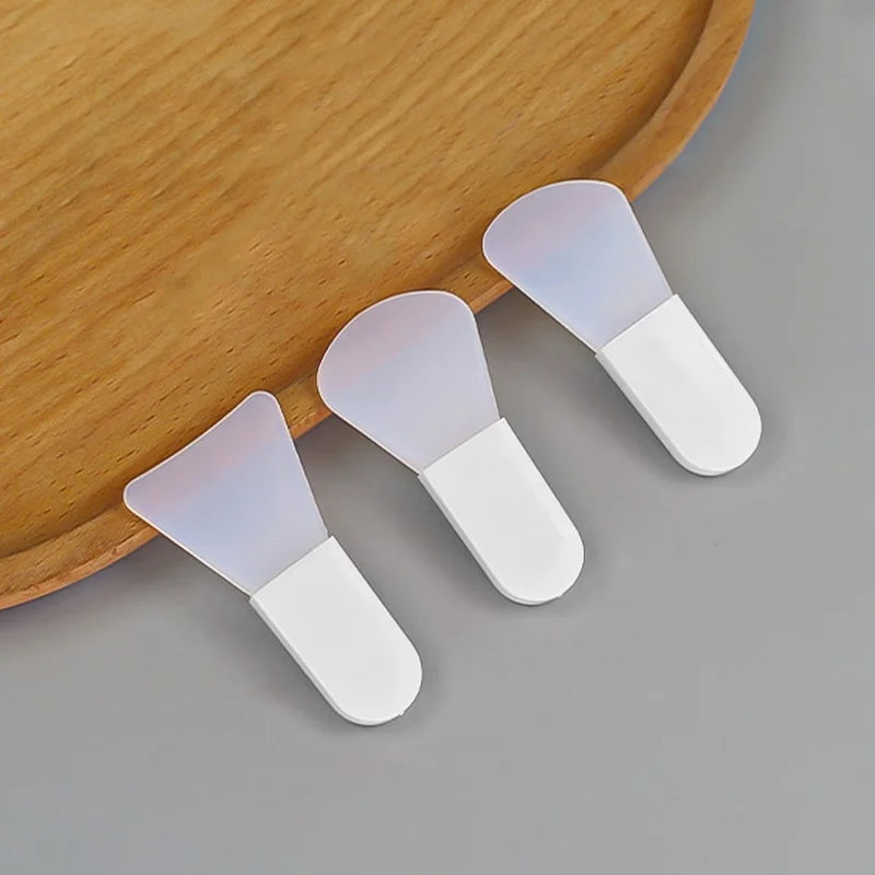 Three white silicone facial mask applicator scrapers with flexible, translucent, fan-shaped tips on a wooden tray.
