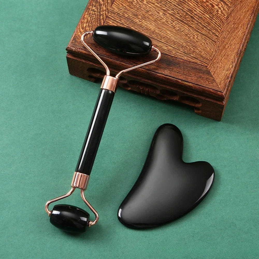 Black jade dual-ended face roller and heart-shaped gua sha stone with gold metal accents on a green background.