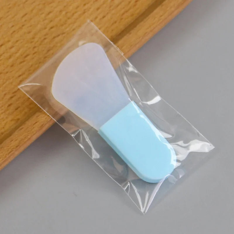 Clear silicone facial mask applicator with a light blue handle, packaged in a small, clear plastic bag.