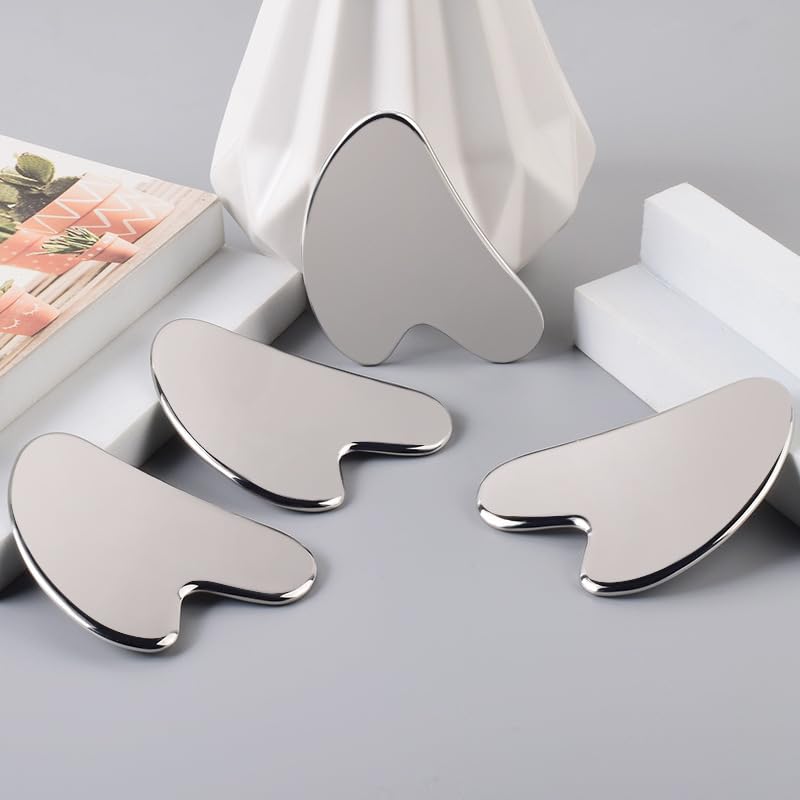 Three smooth, heart-shaped stainless steel Gua Sha facial massage tools arranged on a gray surface with decor.
